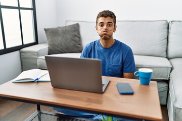 Young handsome hispanic man using laptop sitting on the floor puffing cheeks with funny face. mouth...
