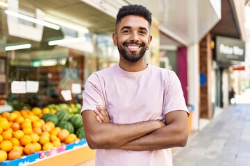 Hispanic man standing by fruits and vegetables shop. Smiling happy with crossed arms by marketplace