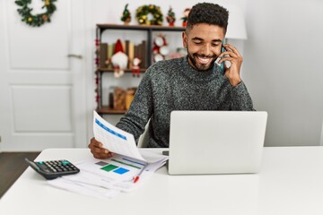 Young african american man controlling economy using laptop and talking on the smartphone at home.