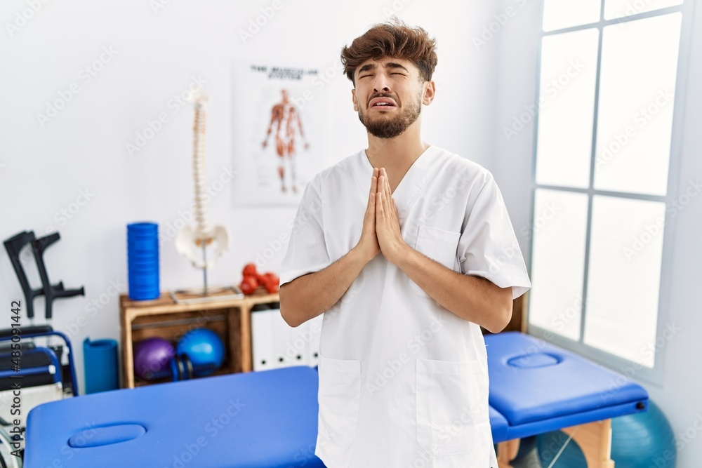 Wall mural Young arab man working at pain recovery clinic begging and praying with hands together with hope expression on face very emotional and worried. begging.