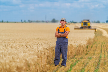 Naklejka na ściany i meble Young attractive farmer standing in wheat field. Combine harvester working in wheat field in background.
