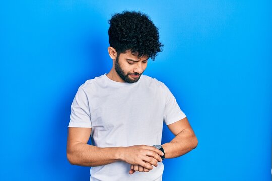 Young arab man with beard wearing casual white t shirt checking the time on wrist watch, relaxed and confident
