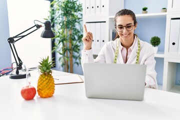 Young hispanic woman working at dietitian clinic smiling happy pointing with hand and finger to the...