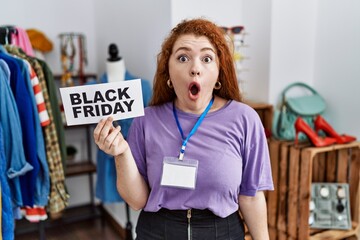 Young redhead woman holding black friday banner at retail shop scared and amazed with open mouth for surprise, disbelief face