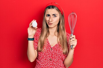 Beautiful hispanic woman holding egg and baker whisk skeptic and nervous, frowning upset because of problem. negative person.