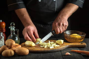 The chef cuts raw potatoes into small pieces with a knife. Close-up of a cook hands while working in kitchen. Cooking delicious breakfast or dinner