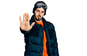 Young hispanic man with beard wearing snow wear and sky glasses doing stop sing with palm of the hand. warning expression with negative and serious gesture on the face.