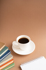 Vertical frame, white cup with a saucer of strong black coffee, books and a notepad on a brown background