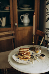 a stack of pancakes on a plate and a jar of jam on a white table. breakfast with pancakes by a burning candle.