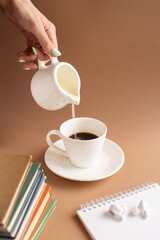 Vertical frame, a white cup with a saucer of strong black coffee, headphones, books and a notepad on a brown background. A woman's hand pours milk.