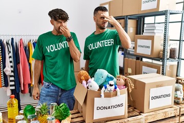 Young gay couple wearing volunteer t shirt at donations stand smelling something stinky and...