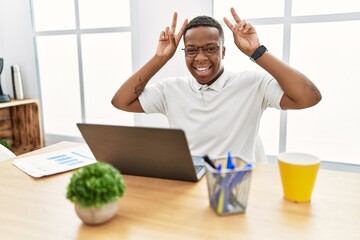 Young african man working at the office using computer laptop posing funny and crazy with fingers on head as bunny ears, smiling cheerful