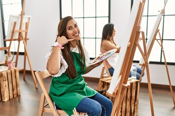 Young hispanic artist women painting on canvas at art studio pointing to the back behind with hand and thumbs up, smiling confident