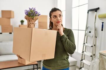 Young hispanic woman holding cardboard box at moving serious face thinking about question with hand...