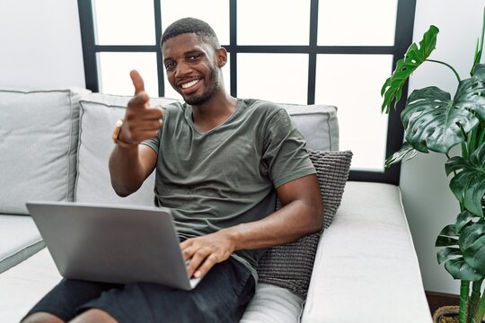 Young african american man using laptop at home sitting on the sofa pointing fingers to camera with happy and funny face. good energy and vibes.