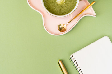 Cute pink cup with heart saucer and matcha latte and golden teaspoon on light green background and notepad with golden pen, girl's morning