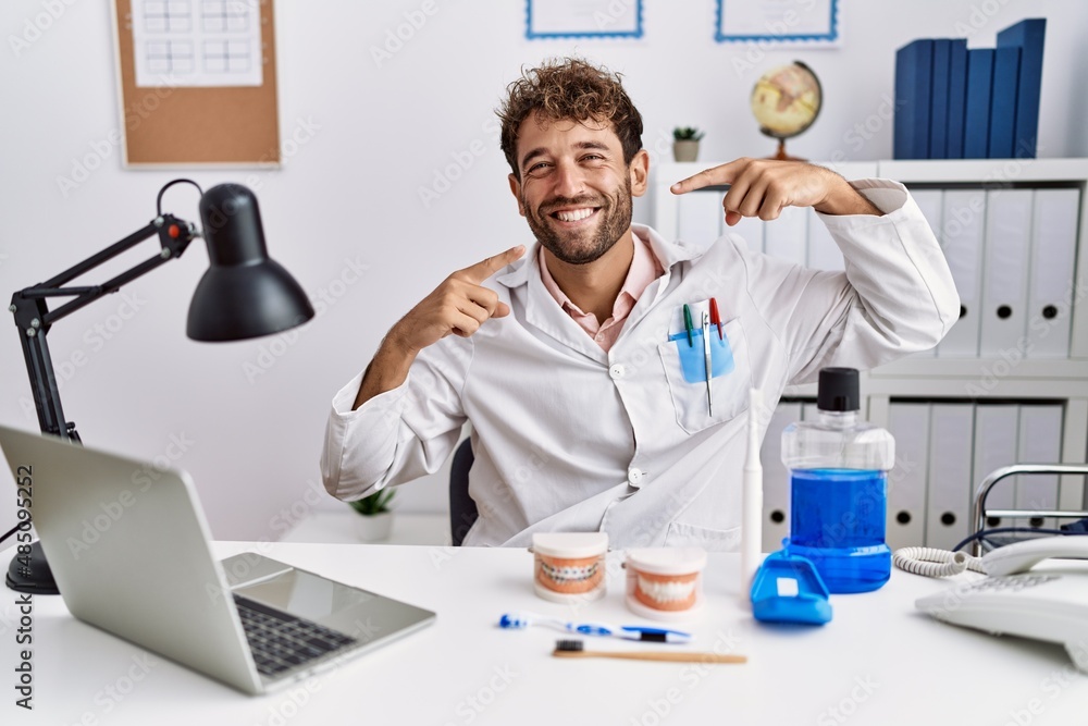 Wall mural Young hispanic dentist man working at medical clinic smiling cheerful showing and pointing with fingers teeth and mouth. dental health concept.