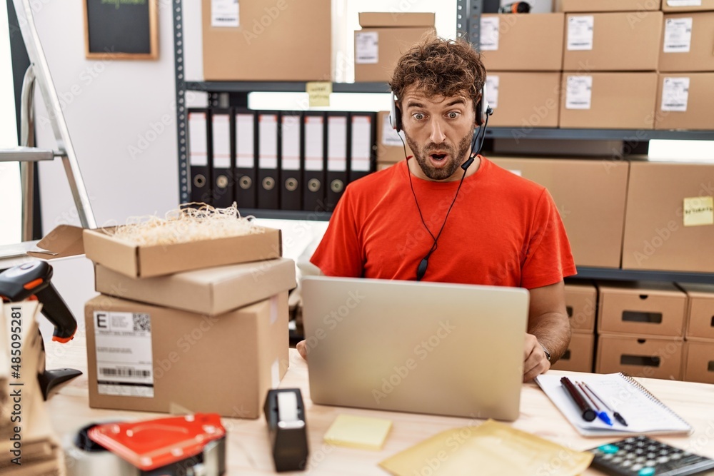 Wall mural Young hispanic call center agent man working at warehouse in shock face, looking skeptical and sarcastic, surprised with open mouth