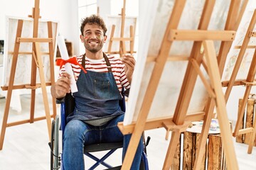 Young hispanic man holing art diploma sitting on wheelchair at art studio