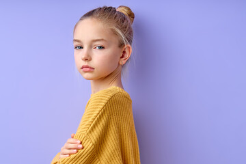 blonde girl looking at camera while standing with arms crossed isolated over purple background. caucasian child in casual yellow shirt is looking gorgeous, little model. children concept - obrazy, fototapety, plakaty