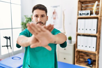 Young physiotherapist man working at pain recovery clinic rejection expression crossing arms and palms doing negative sign, angry face