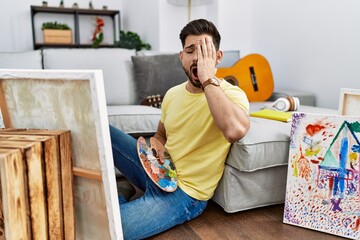 Young man with beard painting canvas at home yawning tired covering half face, eye and mouth with...