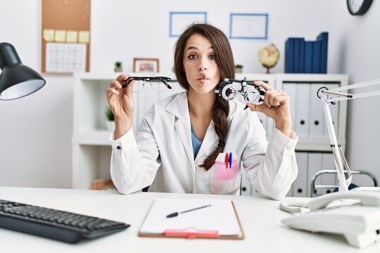Young doctor woman holding optometry glasses and normal glasses making fish face with mouth and squinting eyes, crazy and comical.
