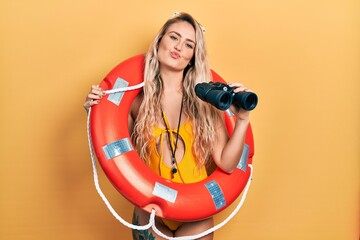 Beautiful young blonde woman wearing bikini and holding lifeguard float and binoculars looking at the camera blowing a kiss being lovely and sexy. love expression.