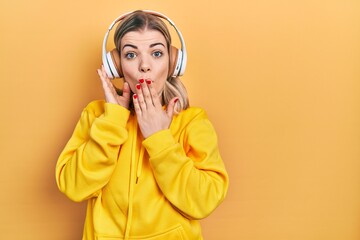 Beautiful caucasian woman listening to music using headphones covering mouth with hand, shocked and...