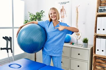 Young caucasian physiotherapist woman holding pilates ball pointing finger to one self smiling...