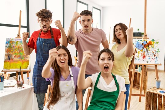 Group of five hispanic artists at art studio angry and mad raising fist frustrated and furious while shouting with anger. rage and aggressive concept.