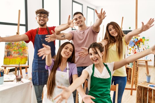 Group of five hispanic artists at art studio looking at the camera smiling with open arms for hug. cheerful expression embracing happiness.