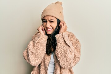 Young hispanic woman wearing wool sweater and winter hat covering ears with fingers with annoyed...