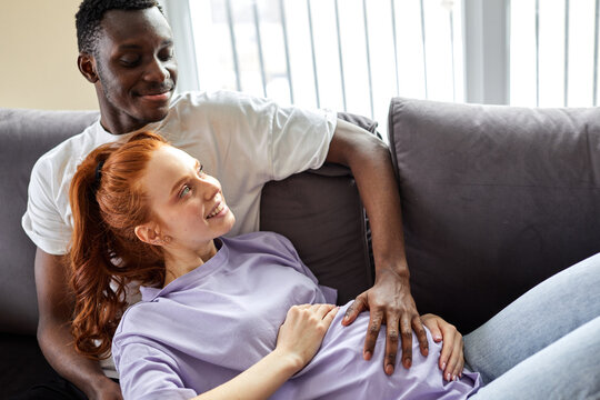 Affectionate male stroking wife's pregnant belly, lying on sofa at home, relaxing. millennial black man and caucasian woman on comfortable couch, dream of bright happy future together in living room