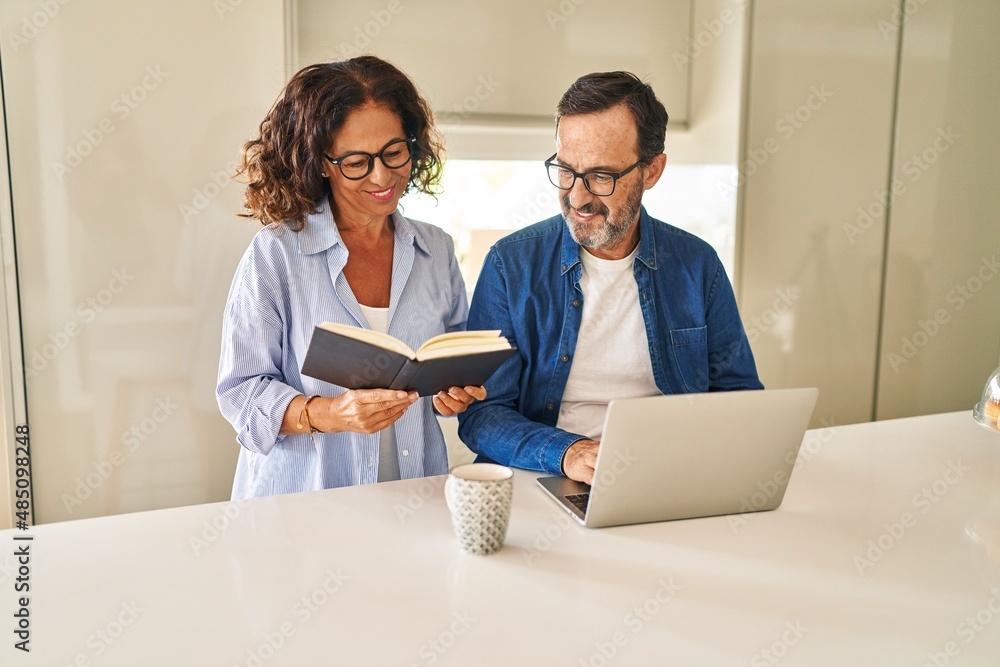 Poster middle age hispanic couple reading book and using laptop at kitchen