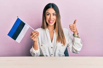Young hispanic woman holding estonia flag sitting on the table smiling happy and positive, thumb up doing excellent and approval sign