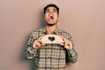 Young african american man holding bread loaf with heart shape angry and mad screaming frustrated and furious, shouting with anger looking up.
