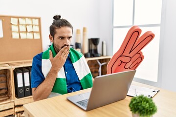 Young hispanic man watching football on laptop cheering game at the office covering mouth with...