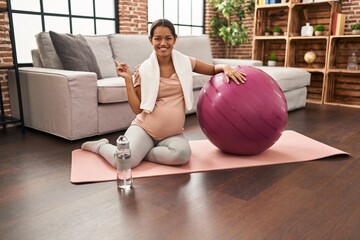 Young pregnant woman sitting on yoga mat with pilates ball smiling happy pointing with hand and...