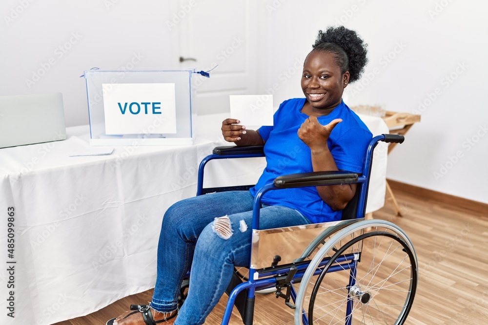 Sticker Young african woman sitting on wheelchair voting putting envelop in ballot box pointing to the back behind with hand and thumbs up, smiling confident