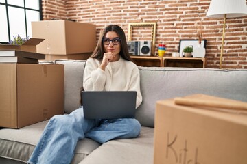 Young hispanic woman sitting on the sofa at new home using laptop clueless and confused with open arms, no idea and doubtful face. - obrazy, fototapety, plakaty
