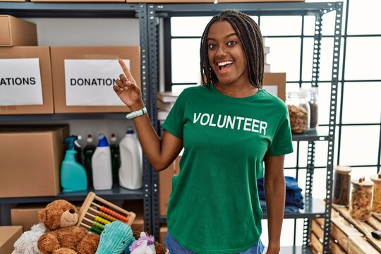 Young african american woman working wearing volunteer t shirt at donations stand with a big smile on face, pointing with hand finger to the side looking at the camera.