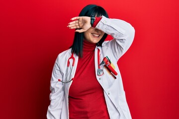 Young hispanic woman wearing doctor uniform and stethoscope covering eyes with arm smiling cheerful and funny. blind concept.