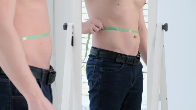 A man measures his waist with a centimeter at the mirror. Slimming and sport, healthy eating concept.