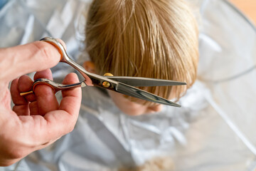 Man's hands doing toddler haircut with hairdresser scissors at home. Baby boy hair cut diy