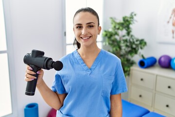 Young physiotherapist woman holding therapy massage gun at wellness center looking positive and happy standing and smiling with a confident smile showing teeth