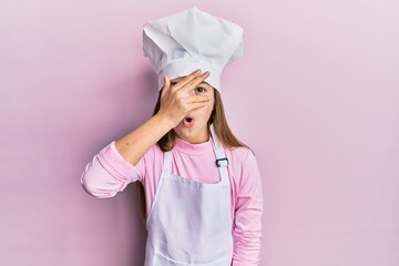 Beautiful brunette little girl wearing professional cook apron and hat peeking in shock covering...