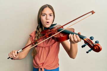 Beautiful brunette little girl playing violin in shock face, looking skeptical and sarcastic, surprised with open mouth