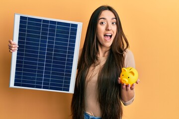 Young hispanic girl holding photovoltaic solar panel and piggy bank celebrating crazy and amazed...