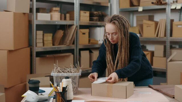 Online store owner. Cute caucasian woman with dreadlocks wearing glasses putting sticker with client address working at the warehouse. Post service and small business concept.